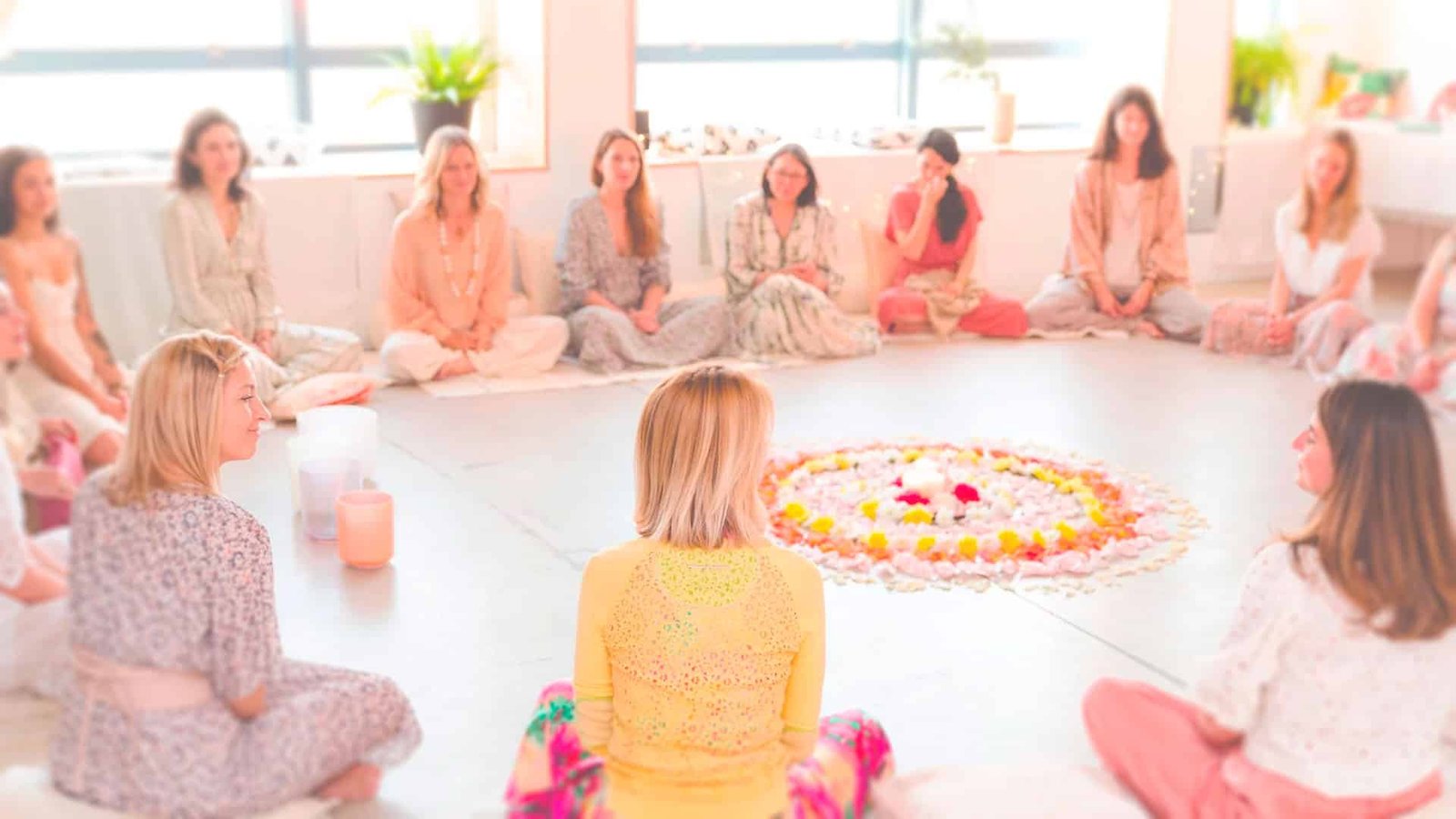Photographie d'un groupe de femmes assises en cercle dans une salle très bien éclairée de lumière naturelle, en train de réaliser un cercle de parole dans la bienveillance. Des bougies et des pétales de fleur sont posés au soleil insistant sur le bien-être et la détente de l'évènement